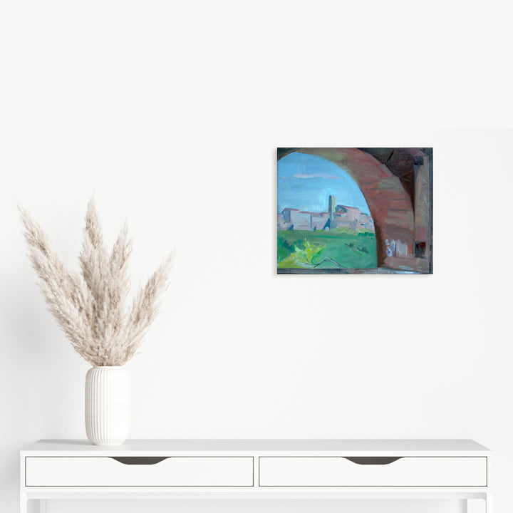 The artwork 'Through the Arch, Casole' by Peter Colquhoun captures a scenic Italian landscape viewed through a terracotta stone archway. The composition features vibrant blue skies, distant medieval buildings on a hilltop, and lush green countryside, all framed by the rustic arch's curved stonework. Oil on linen, 13x16 inches.