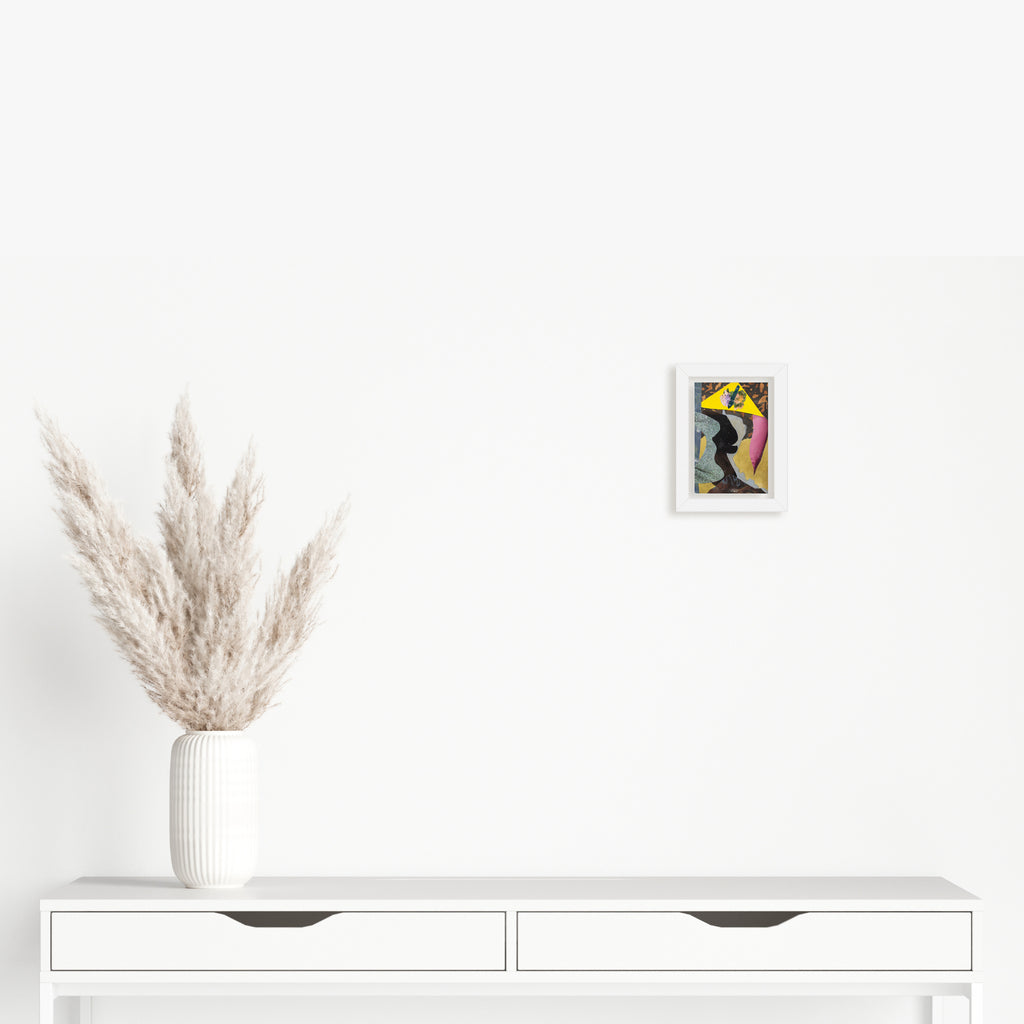 The artwork 'Fragment#14' by Winnie Sidharta features a small abstract mixed media composition displayed on a white wall above a minimalist console table. The artwork combines watercolor and magazine cutouts on panel, showcasing vibrant yellow and dark tones in a 4x6 inch format. A white vase with fluffy pampas grass decorates the scene by Winnie Sidharta.
