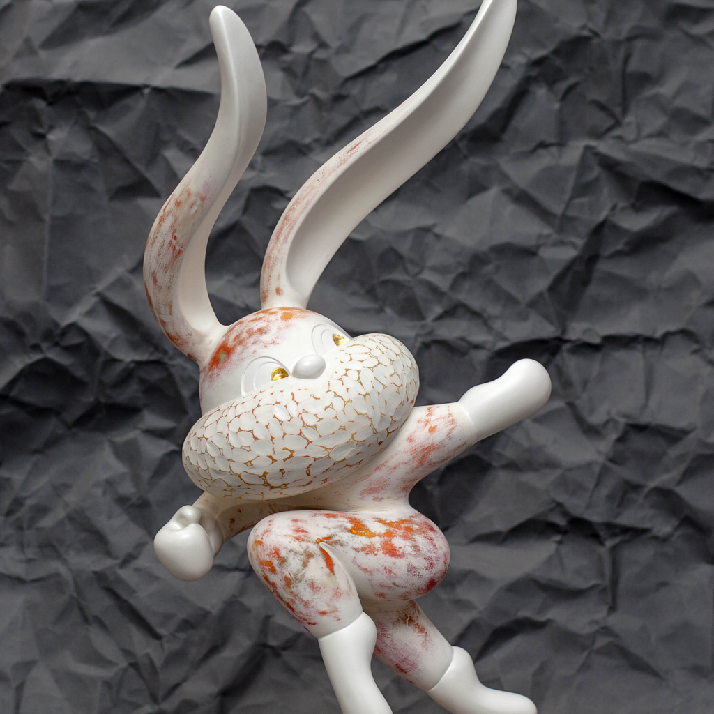 The artwork 'HOPE SPRINGS ETERNAL series' by Yusuke Ochiai features a dynamic white resin sculpture depicting an abstract bunny figure in motion. The piece showcases intricate crackle patterns adorned with coral-orange accents and 22k gold leaf details, set against a textured dark background. The sculpture combines smooth curves with delicate surface treatments.