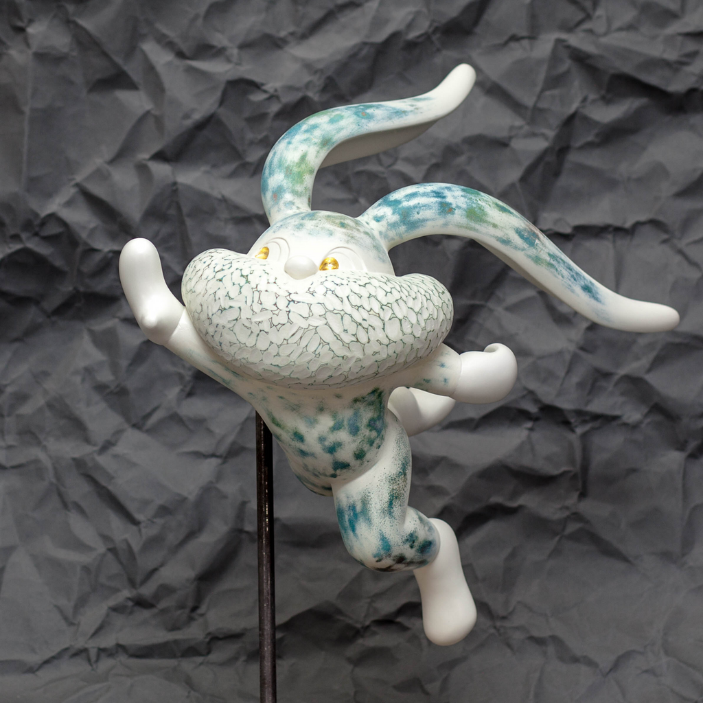 The artwork 'HOPE SPRINGS ETERNAL series' by Yusuke Ochiai features a whimsical resin sculpture of a playful creature in white with turquoise speckled patterns. The figure displays dynamic movement, with curved limbs and textured body details enhanced by spray paint and 22k gold leaf accents against a dark background. A unique contemporary art piece mounted on a stand.