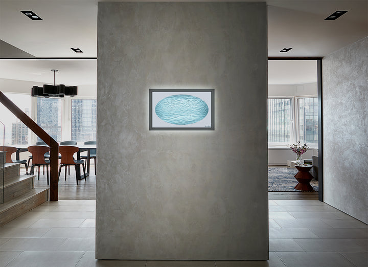 The artwork 'Emptiness.here.2020 #1' by Daniel Kersh features an illuminated oval-shaped artwork in ethereal aqua blue tones mounted on a concrete-textured gray wall. The piece combines oil paint and giclee printing on canvas, creating a serene rippling effect, displayed in a modern interior setting with a custom floating frame and integrated lighting element by Daniel Kersh.