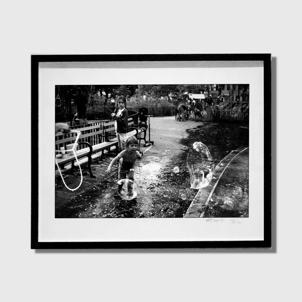 The artwork 'Bubble Catastrophe' captures a nostalgic black and white scene in a park where a person playfully interacts with soap bubbles against a backdrop of benches and trees. Water reflections and dramatic shadows create depth in this 32x40 inch fine art photograph. By Mari Sarai.