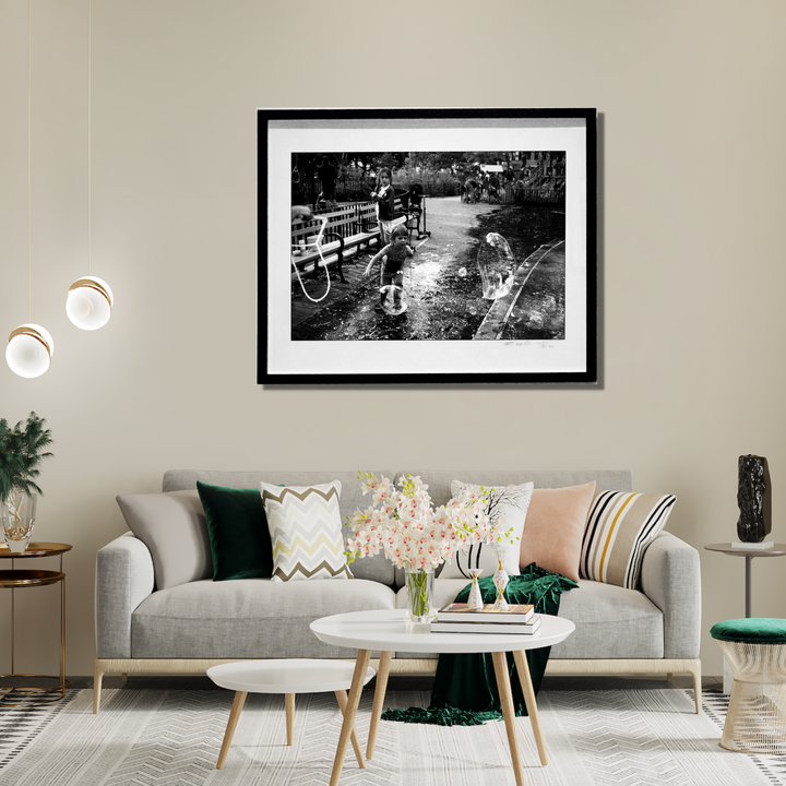 The artwork 'Bubble Catastrophe' by Mari Sarai features a nostalgic black and white photograph showing children at play with soap bubbles in a park setting. The composition captures playful moments around metal benches and a paved pathway, creating a timeless scene of childhood joy. The framed monochromatic print, measuring 32x40 inches, showcases natural lighting and documentary-style photography. Edition 20 by Mari Sarai.