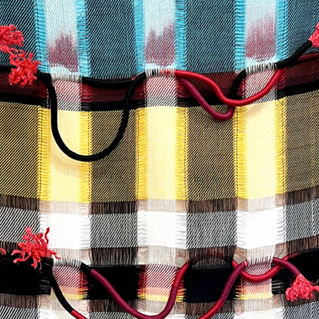 The artwork 'Resisting The Fire' by Kathie Halfin features a bold plaid pattern in turquoise, yellow, white, and black, interwoven with red rope accents. The textile piece showcases intricate weaving techniques combining cotton and linen, with copper wire and nylon rope creating dimensional elements against the geometric background.