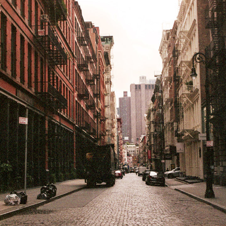 The artwork 'Ghost Neighborhood in Soho' by Mari Sarai captures a moody urban street scene featuring historic cast-iron buildings with fire escapes, cobblestone streets, and distant high-rises in New York's Soho district. Shot in muted sepia tones, the composition creates depth through converging lines of architecture, parked vehicles, and characteristic iron balconies. 40x32 inches, Edition 20. By Mari Sarai.