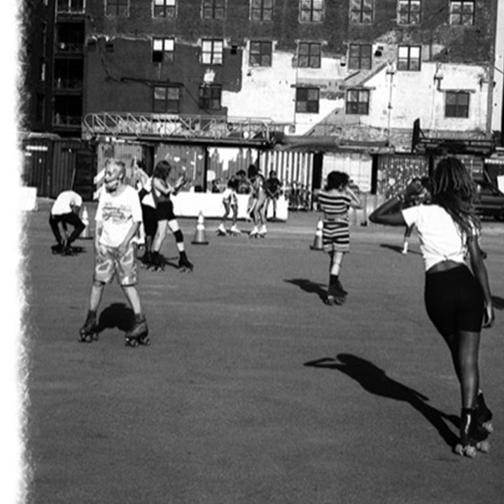 The artwork 'Roller Wave' captures a dynamic black and white urban scene showing people roller skating in an outdoor rink against a backdrop of apartment buildings. The composition features strong shadows cast on the concrete surface, creating geometric patterns that complement the movement of the skaters. The monochromatic treatment emphasizes the nostalgic, documentary-style photography. By Mari Sarai.