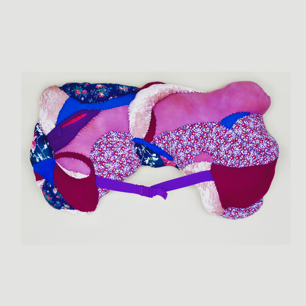 The artwork 'Clavicle' by Hanna Washburn features abstract organic shapes crafted from recycled textiles in vibrant pink, royal blue, and burgundy hues. Floral patterns and soft textures combine with plush white accents, secured by a purple seatbelt strap, mounted on a wooden frame. The sculptural piece evokes anatomical forms through quilted fabric composition.