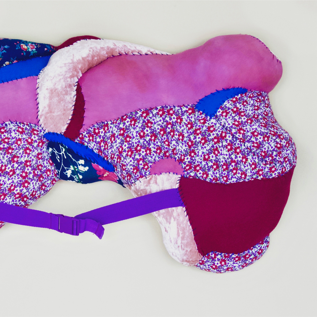 The artwork 'Clavicle' by Hanna Washburn features a sculptural textile piece combining vibrant purple, pink, and blue floral patterns with plush white accents. The abstract form curves organically, secured with a purple seatbelt strap, showcasing recycled fabrics and textiles meticulously stitched together in a padded, three-dimensional composition. By Hanna Washburn.