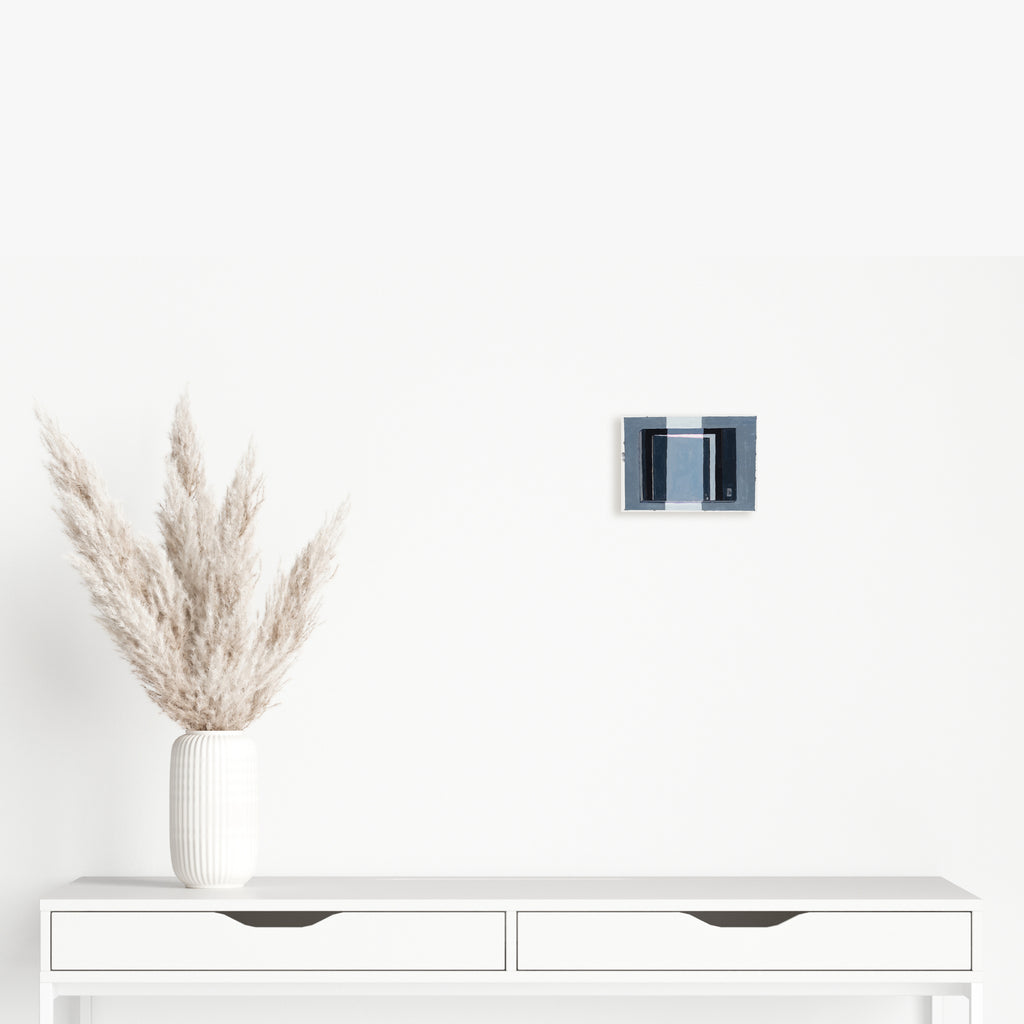 The artwork 'Closing Door 2' by Til Will features a small black and indigo abstract painting depicting a geometric doorway or window frame against a white background. Mounted above a minimalist white console table with pampas grass in a ribbed vase, the 5x7 inch acrylic on paper piece creates a striking architectural focal point.