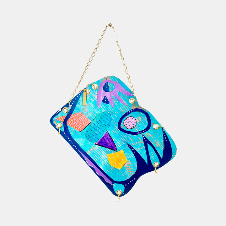 The artwork 'Coastal' by Sunny Chapman features vibrant geometric shapes on a turquoise background. Abstract triangles in purple, yellow, and coral float alongside circular patterns, creating a playful and modern composition. The acrylic on paper piece incorporates navy blue curved elements and delicate dotted details, suspended by a gold chain, measuring 16x12 inches by Sunny Chapman.