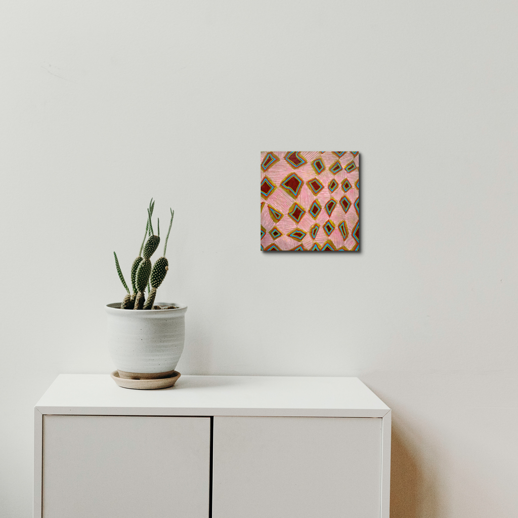 The artwork 'Composition No. 235' by Raul Ortiz features a geometric abstract pattern of repeating diamonds in pink and brown tones on linen canvas, displayed above a white cabinet with a potted cactus. The 12x12 inch acrylic painting creates a rhythmic, modernist design with precise brushwork and balanced composition by Raul Ortiz.