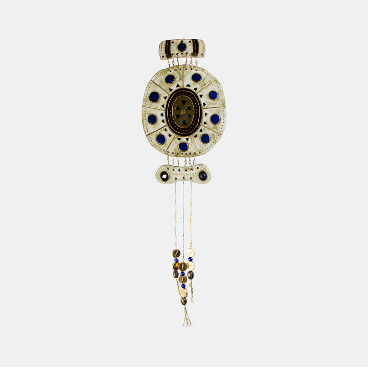 The artwork 'December' by Adina Andrus features an ornate oval pendant design in glazed stoneware with deep blue cabochons arranged in a symmetrical pattern. The central medallion displays intricate 24k gold detailing, surrounded by a cream-colored ceramic frame. Delicate metal chains with decorative beads cascade from the bottom, creating an elegant hanging sculpture.