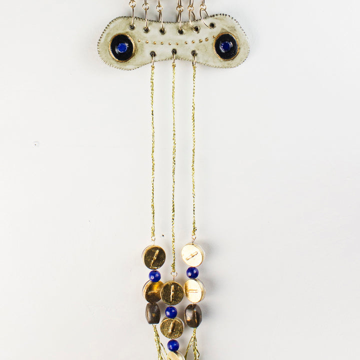 The artwork 'December' by Adina Andrus features a sculptural wall piece with dual sapphire-blue cabochons set in a curved cream-colored ceramic form, adorned with delicate gold chains suspending amber and ivory glass beads. The 36-inch stoneware creation combines glazed ceramics, metalwork, and 24k gold accents in an elegant vertical composition.