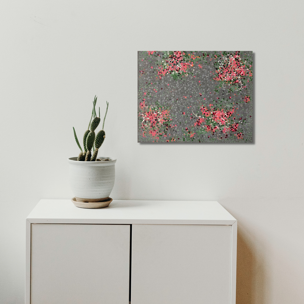 The artwork 'Desert Bloom' by Shira Toren features delicate clusters of coral pink and sage green dots scattered across a textured gray backdrop, created with Venetian plaster and pigments on canvas. The organic arrangement suggests flowering desert plants, with swirling patterns reminiscent of wind-swept blooms.
