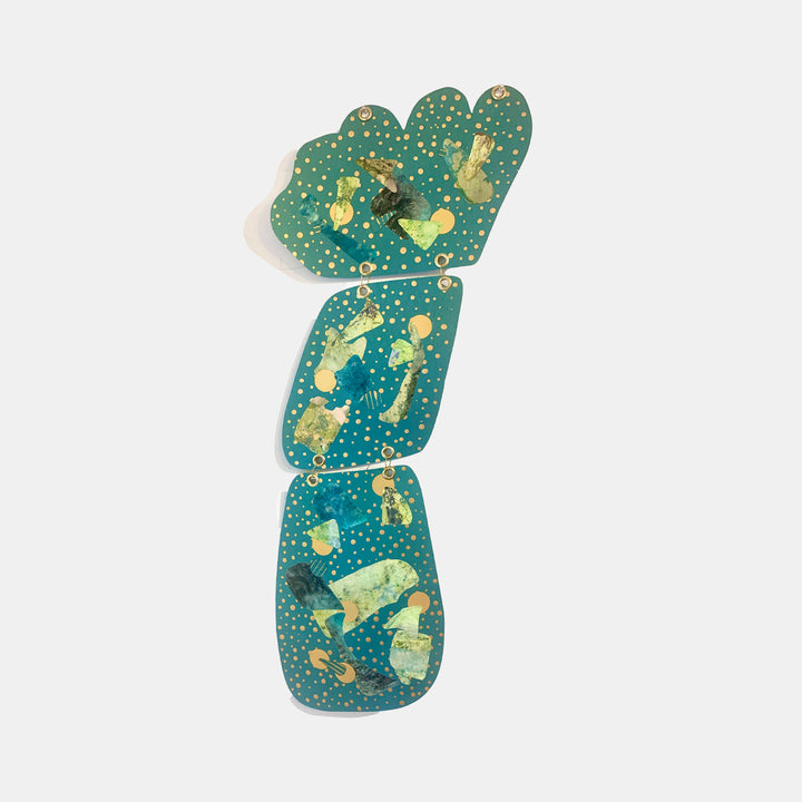 The artwork 'Dotter' by Sunny Chapman features three connected geometric shapes in teal with gold metallic accents. Abstract collage elements and dotted patterns create a cosmic effect on metallic paper. The vertical composition measures 40x12 inches, with grommets connecting the shapes into a dynamic wall hanging.