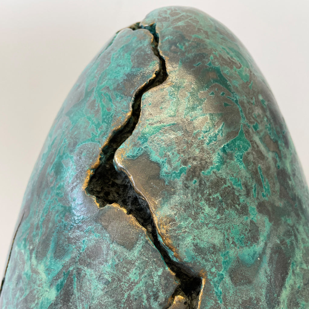 The artwork 'EGG' by Nora Chavooshian features a sculptural form in cast forton with striking verdigris bronze patina. A dramatic crack bisects the oval shape, revealing dark depths within. The surface showcases a mesmerizing interplay of mottled turquoise and aged metallic bronze textures, creating an organic, weathered appearance.