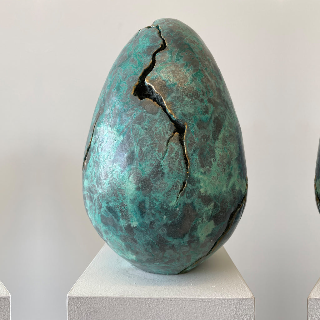 The artwork 'EGG' by Nora Chavooshian features a large sculptural egg form with a striking verdigris bronze patina. A dramatic jagged crack splits the surface, creating organic textural patterns. The cast forton sculpture, measuring 12 inches tall, showcases a rich turquoise-green oxidized finish that mimics ancient bronze artifacts.