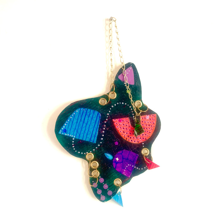 The artwork 'Feed' features abstract shapes in vibrant colors of emerald green, electric blue, hot pink, and deep purple on a pendant-like form. Bold geometric patterns and dotted textures create a dynamic composition, with metallic gold accents and a delicate chain. This 12x12 inch acrylic on paper piece showcases a playful, modern aesthetic by Sunny Chapman.