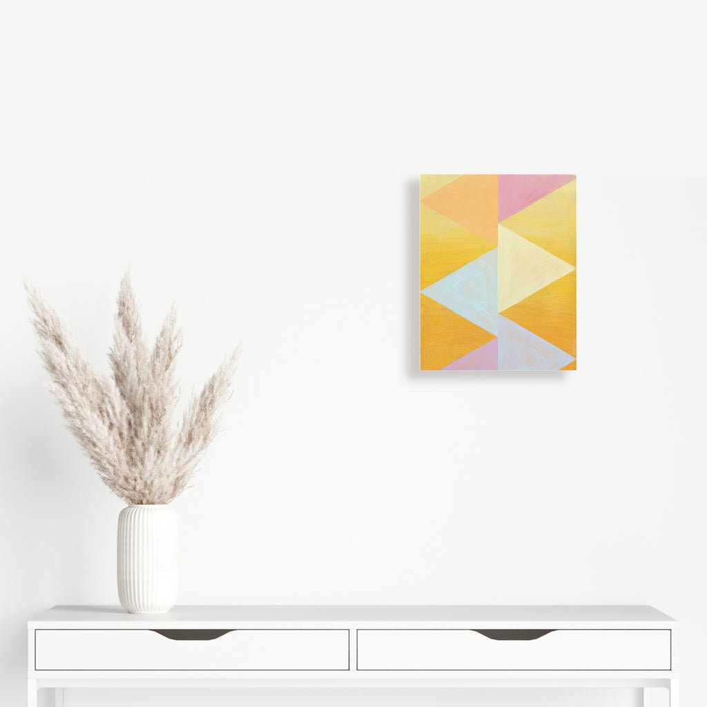 The artwork 'Flag Me' by Amber Heaton features geometric triangular shapes in soft pastel colors including yellow, pink, orange, and mint blue arranged in a modern abstract pattern on wood. Acrylic paint creates clean lines and color blocks, measuring 15x12 inches, displayed above a white console with decorative pampas grass.