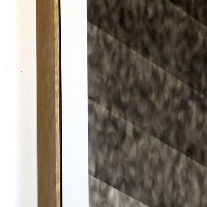The artwork 'Foot Habits' by Evan Ishmael features a minimalist composition with diagonal lines creating a striking contrast between light and dark. A textured, smoky gray pattern created with soot and spray-applied acrylic spans across the canvas, while a clean white vertical stripe and wooden frame element adds structural balance to the contemporary abstract piece.