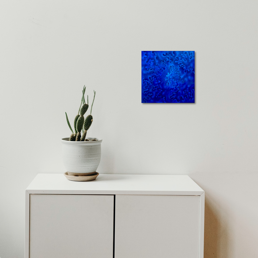 The artwork 'Fury of the Seasons' by Joe Piscopia features a vibrant abstract composition in deep electric blue tones. The painting showcases a radiating pattern from the center, with swirling textures creating a dynamic, storm-like effect. The oil on canvas piece glows against a minimalist white wall, displayed above a white cabinet with a potted cactus.