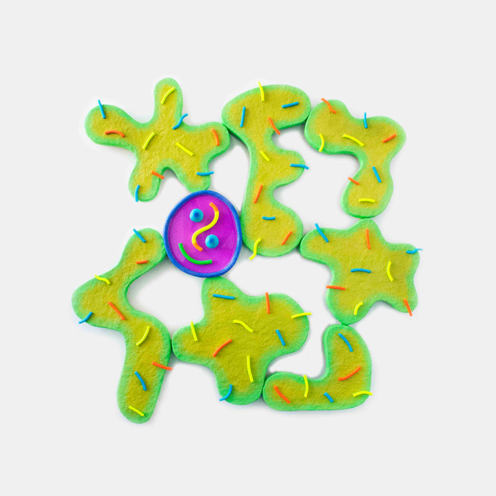 The artwork 'Good Germs' by Ryan Patrick Martin features abstract organic shapes in vibrant yellow-green tones surrounding a central purple resin circle with a playful smiley face. The sculptural piece combines wood, epoxy, and acrylic paint, creating a whimsical, biomorphic composition against a white background.