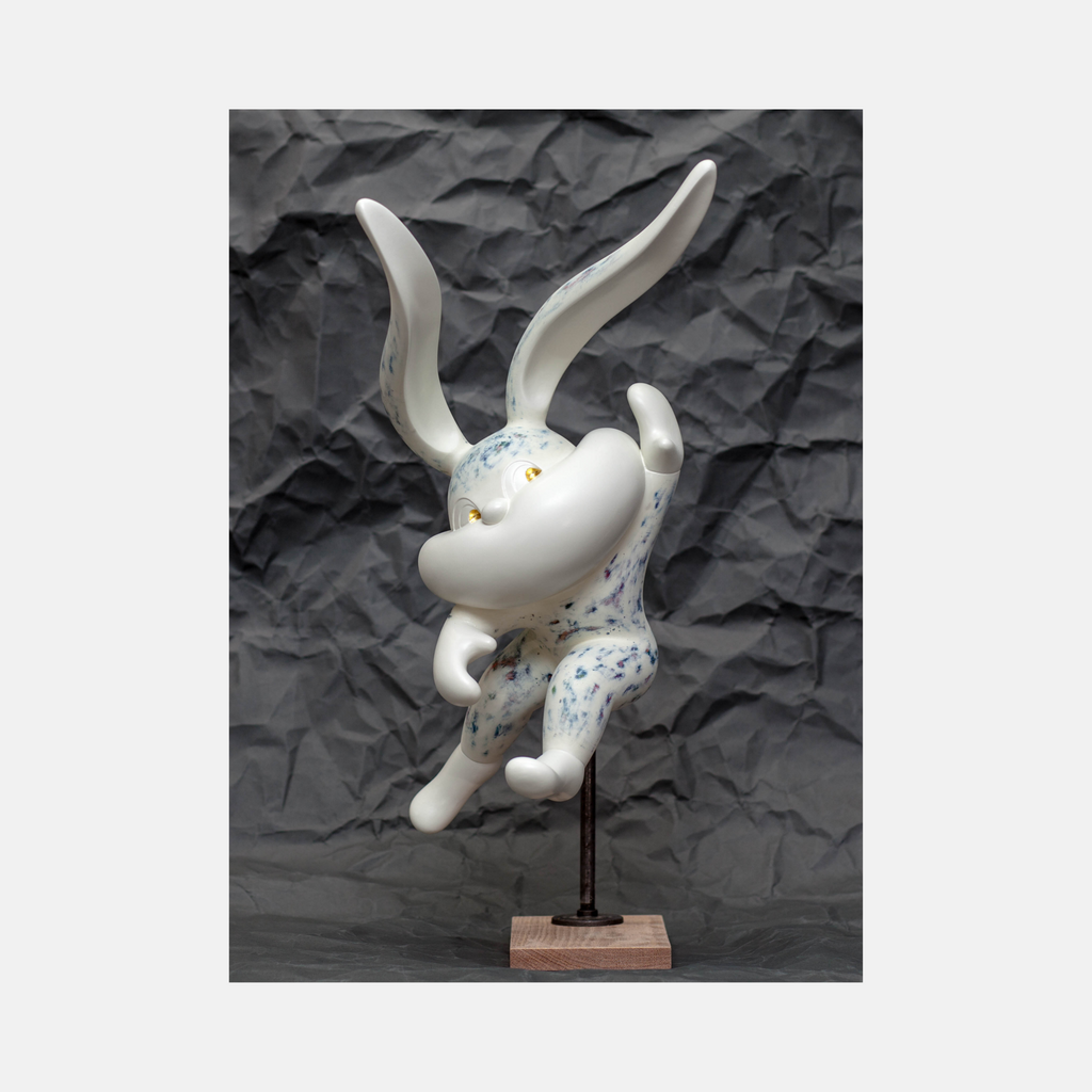 The artwork 'HOPE SPRINGS ETERNAL series' by Yusuke Ochiai features a whimsical white resin rabbit sculpture with elongated ears, adorned with delicate blue speckled patterns and 22k gold leaf accents. The playful figure appears to be leaping upward, mounted on a wooden base against a textured dark background.
