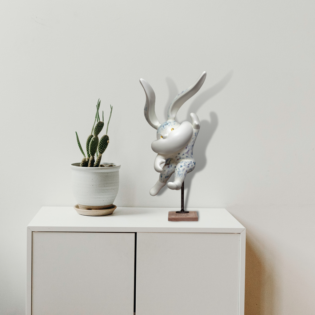 HOPE SPRINGS ETERNAL series by Yusuke Ochiai: A contemporary sculpture featuring a whimsical white rabbit figure with elongated ears in resin, adorned with gold leaf details and spray finish, mounted on a stand. Displayed alongside a potted cactus on a white minimalist cabinet, creating a modern artistic composition.