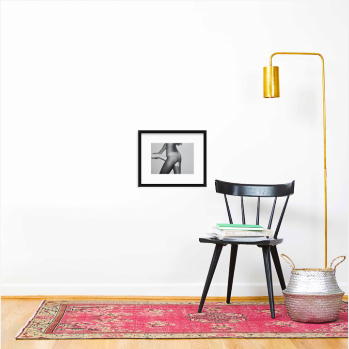 The artwork 'Hand in Between Legs' by Mari Sarai, displayed in a black frame with white matting on a white wall. Modern interior setting features a black wooden chair, brass floor lamp, and vintage pink rug. Clean minimalist composition with a metallic basket as accent. Framed photograph measures 8.5x11 inches. By Mari Sarai.