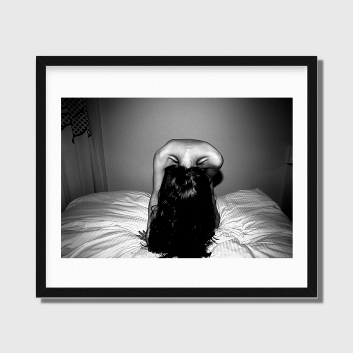 The artwork 'Headbanging' by Mari Sarai captures a dramatic black and white photograph of flowing dark hair against white bedding. The monochromatic composition features strong contrast and movement, presented in a sleek black frame with white matting. The artistic perspective creates a powerful abstract form through the arched position and cascading hair.