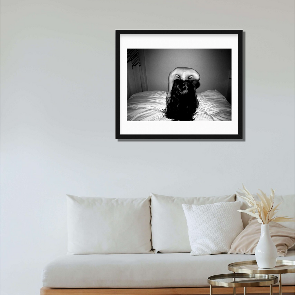 The artwork 'Headbanging' by Mari Sarai features a dramatic black and white photograph of a figure on a bed, captured in an expressive pose with long dark hair in motion. The monochromatic composition creates a striking contrast between light and shadow, presented in a black frame with white matting, measuring 33x25 inches.