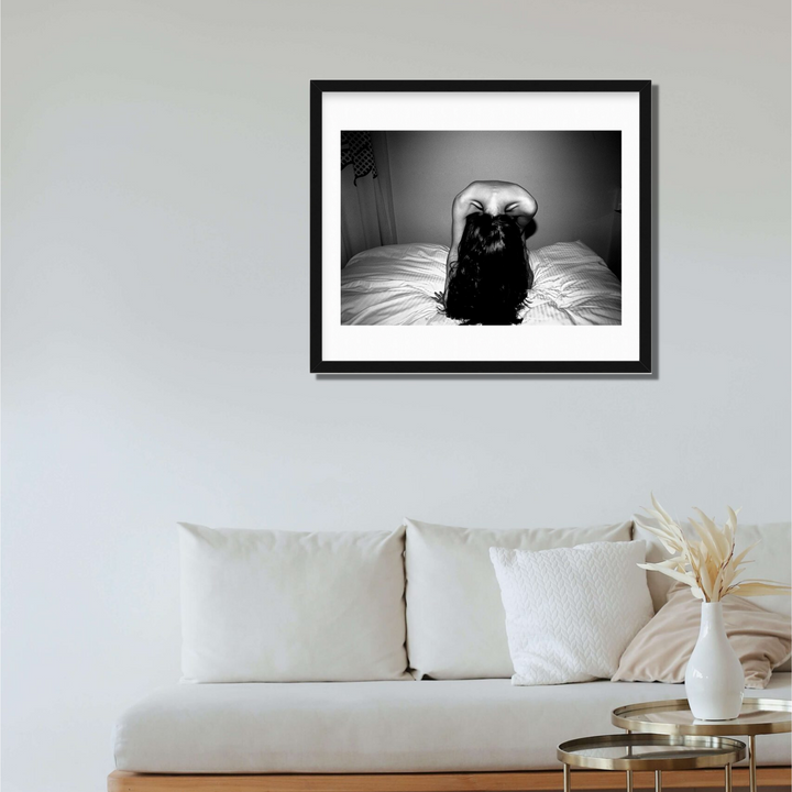 The artwork 'Headbanging' by Mari Sarai features a dramatic black and white photograph of a figure on a bed, captured in an expressive pose with long dark hair in motion. The monochromatic composition creates a striking contrast between light and shadow, presented in a black frame with white matting, measuring 33x25 inches.