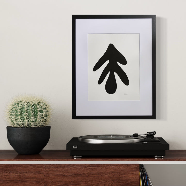 The artwork 'Morning Meditation XIV' by Shadia Sabagh features a bold black abstract leaf-like shape on white paper, framed in black with white matting. Displayed alongside a turntable and potted cactus, this minimalist acrylic marker piece creates a striking modern composition. 20x16 inches, mixed media artwork by Shadia Sabagh.