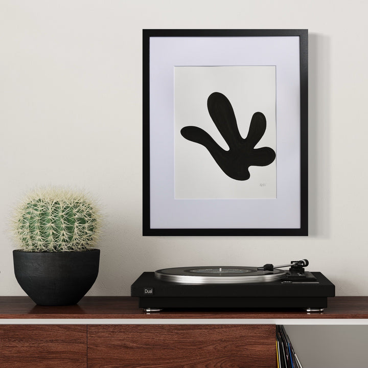 Morning Meditation XIII by Shadia Sabagh features a bold black abstract organic shape resembling a floating leaf or hand gesture against a white background, framed in black with white matting. The minimalist artwork is displayed above a turntable, alongside a potted cactus, creating a modern, zen-like aesthetic.