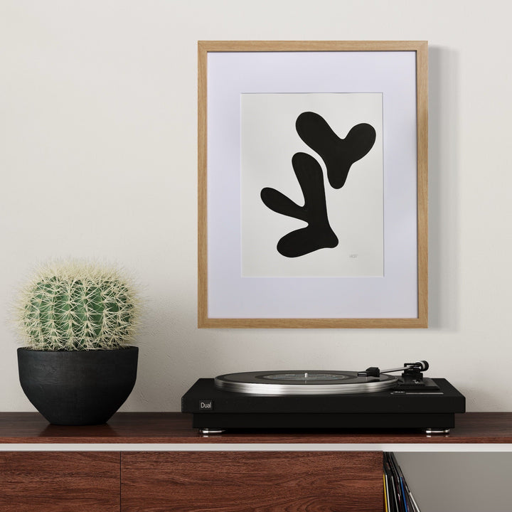 Morning Meditation XII by Shadia Sabagh features a minimalist black abstract form resembling organic shapes against a white background, framed in light wood. Displayed above a turntable alongside a potted cactus, this acrylic marker artwork creates a striking modern composition with flowing, biomorphic lines.