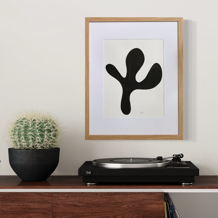 The artwork 'MORNING MEDITATION XI' by Shadia Sabagh features a bold black organic shape resembling a minimalist plant form against white paper, framed in natural wood. Displayed alongside a potted cactus and turntable on wooden furniture, the abstract design evokes a sense of calm and growth. Created with acrylic marker on mixed media paper.