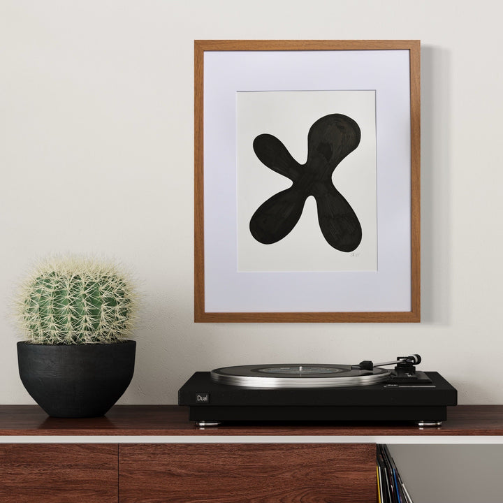 The artwork 'Morning Meditation VIII' by Shadia Sabagh features a bold black organic shape resembling a four-petal flower or abstract cross on white paper, framed in wood. Displayed above a modern record player and beside a potted cactus, the minimalist design creates a striking contrast in a contemporary home setting.