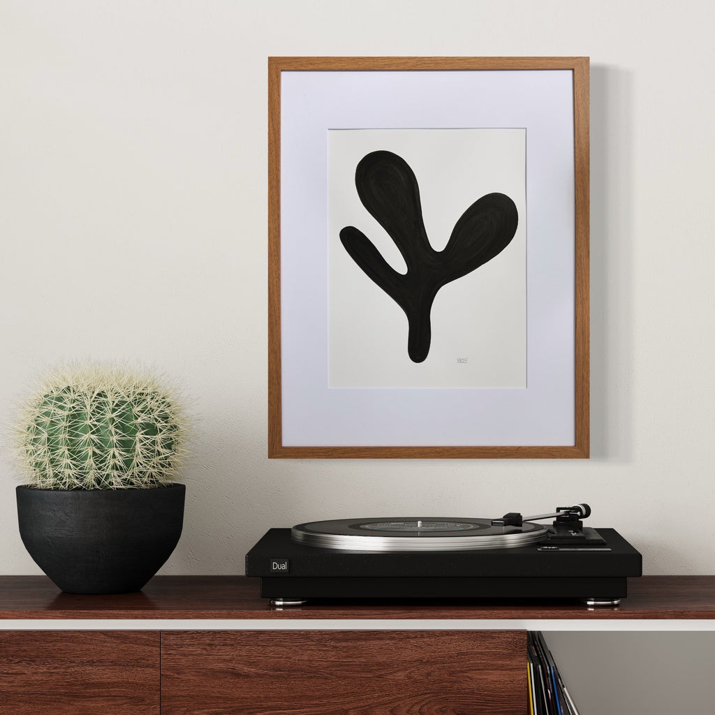 The artwork 'Morning Meditation VII' by Shadia Sabagh features a bold black organic shape resembling a minimalist plant form against a white background, framed in wood and white matting. Displayed alongside a cactus plant and turntable, the abstract composition creates a striking modern aesthetic. Created with acrylic marker on mixed media paper.