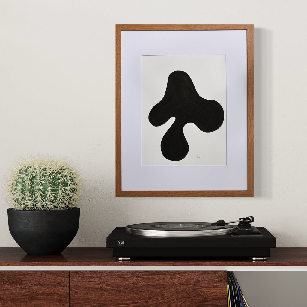 The artwork 'Morning Meditation VI' by Shadia Sabagh features a bold, abstract black organic shape against a white background. The form resembles a fluid, three-pronged silhouette, creating a striking minimalist composition. Presented in a wooden frame with white matting, this acrylic marker piece exemplifies contemporary abstract expressionism.