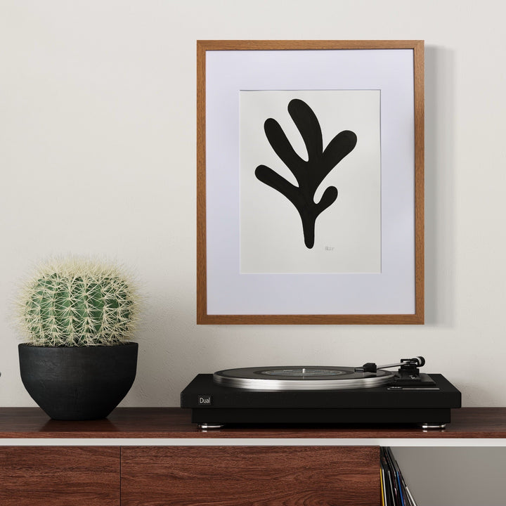 The artwork 'Morning Meditation V' by Shadia Sabagh features a bold black organic shape resembling a stylized leaf or branch against a white background, framed in warm wood. The minimalist composition in acrylic marker showcases fluid, abstract lines forming a botanical-inspired silhouette. Displayed alongside a potted cactus and turntable on wooden furniture.