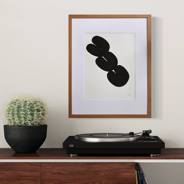 Morning Meditation IV by Shadia Sabagh features a minimalist abstract composition with three interconnected black organic shapes on white paper, framed in wood. The bold, flowing forms create a sense of movement and balance, showcased alongside a cactus plant and turntable in a modern interior setting.