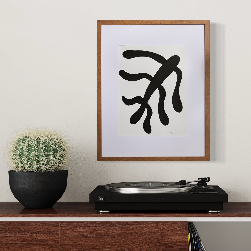 The artwork 'Morning Meditation III' by Shadia Sabagh features a bold black abstract form resembling flowing seaweed or organic branches against a white background, framed in brown wood. The minimalist design creates a striking contrast, displayed alongside a potted cactus and record player on a wooden surface. Created with acrylic marker on mixed media paper.