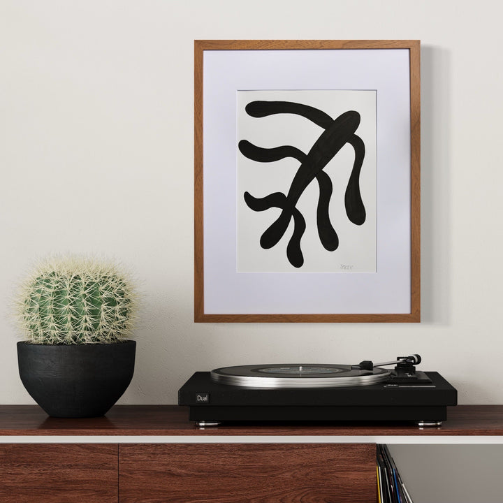 The artwork 'Morning Meditation III' by Shadia Sabagh features a bold black abstract form resembling flowing seaweed or organic branches against a white background, framed in brown wood. The minimalist design creates a striking contrast, displayed alongside a potted cactus and record player on a wooden surface. Created with acrylic marker on mixed media paper.
