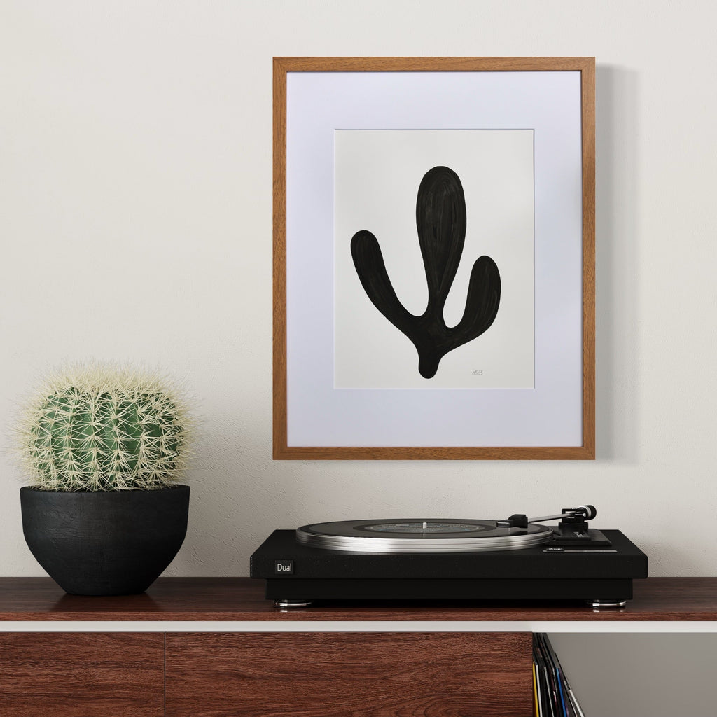 Morning Meditation II by Shadia Sabagh features a bold black organic shape against white background, reminiscent of a minimalist cactus or abstract botanical form. The artwork, framed in wood with white matting, presents a striking monochromatic composition created with acrylic marker on mixed media paper. The simple yet powerful design evokes zen-like tranquility.