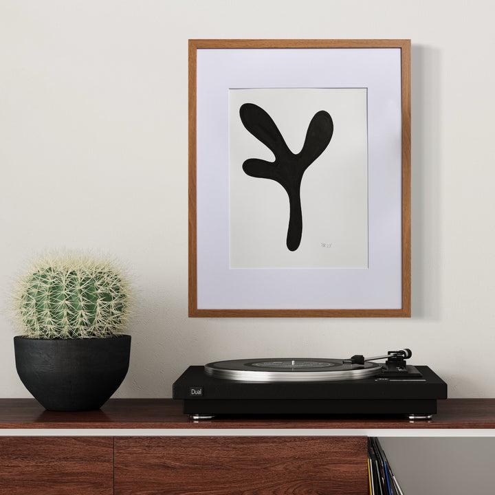The artwork 'Morning Meditation I' by Shadia Sabagh features a bold black organic shape on white paper, resembling a minimalist abstract plant form with curved branches. The piece is elegantly framed in wood with a white mat, displayed alongside a potted cactus and turntable, creating a modern aesthetic composition.