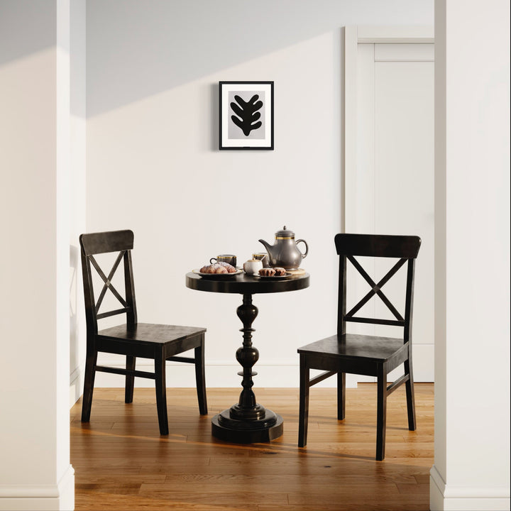 The artwork 'Morning Meditation XXVI' by Shadia Sabagh captures a serene tea setting with two black cross-back chairs flanking an ornate pedestal table. A minimalist black leaf print artwork hangs above on the white wall. The scene features a silver teapot and teacups on the round table, set against warm hardwood flooring and natural lighting.
