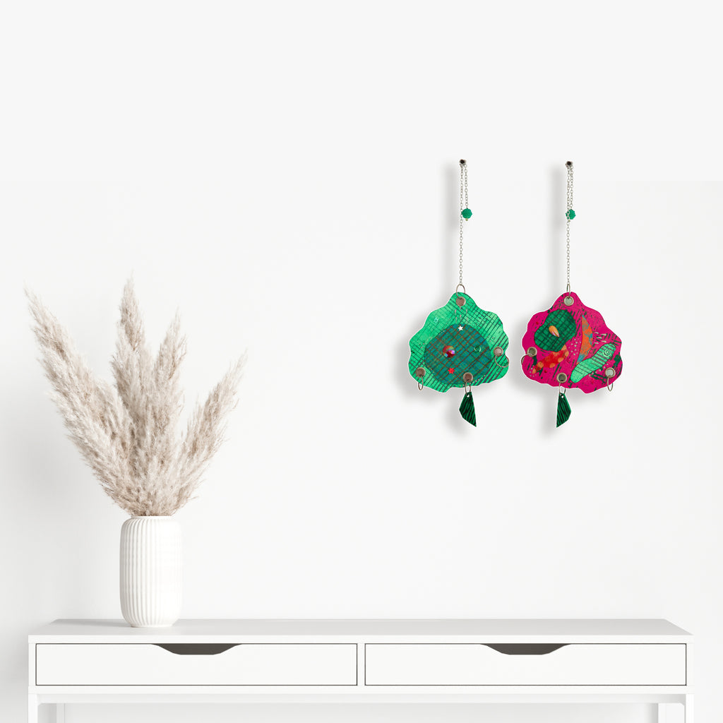 The artwork 'Cloud' by Sunny Chapman features whimsical hanging cloud-shaped decorations in vibrant emerald green and hot pink, adorned with metallic chains, glitter, sequins, and jewels. Mixed media elements include grommets, beads, and handmade charms, creating a playful and luxurious aesthetic with a modern twist.