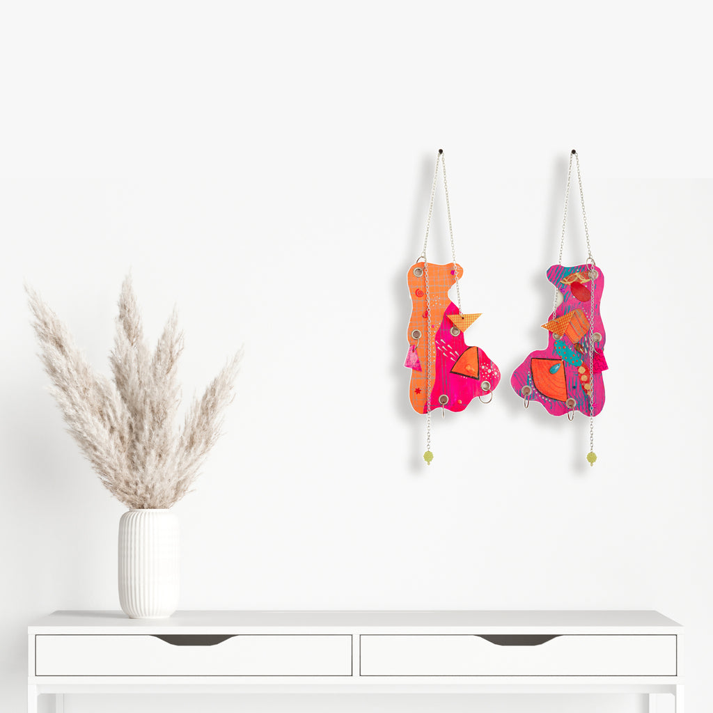 The artwork 'Duck Face' features two vibrant abstract wall hangings in orange and hot pink tones, adorned with glitter, sequins, and metallic accents. Each piece displays playful, curved shapes suspended by silver chains and grommets, complemented by decorative beads and handmade charms. By Sunny Chapman.