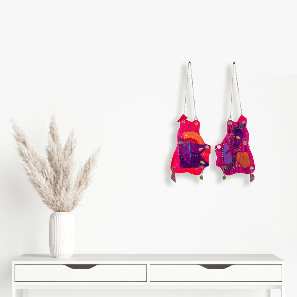 The artwork 'Frog Mouth' by Sunny Chapman features two vibrant hanging pieces in hot pink and purple tones, adorned with glitter, jewels, and metallic accents. Each piece displays an abstract frog-like shape decorated with sequins, chains, and handmade charms, mounted on heavy paper with grommets and metal rings for wall display.