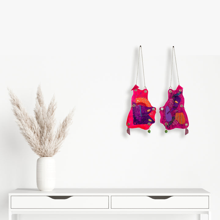 The artwork 'Frog Mouth' by Sunny Chapman features two vibrant hanging pieces in hot pink and purple tones, adorned with glitter, jewels, and metallic accents. Each piece displays an abstract frog-like shape decorated with sequins, chains, and handmade charms, mounted on heavy paper with grommets and metal rings for wall display.